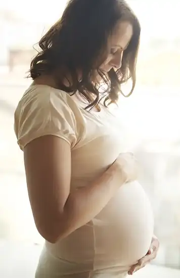 Eine Schwangere hält lächelnd ihren Bauch im hellen Gegenlicht.
